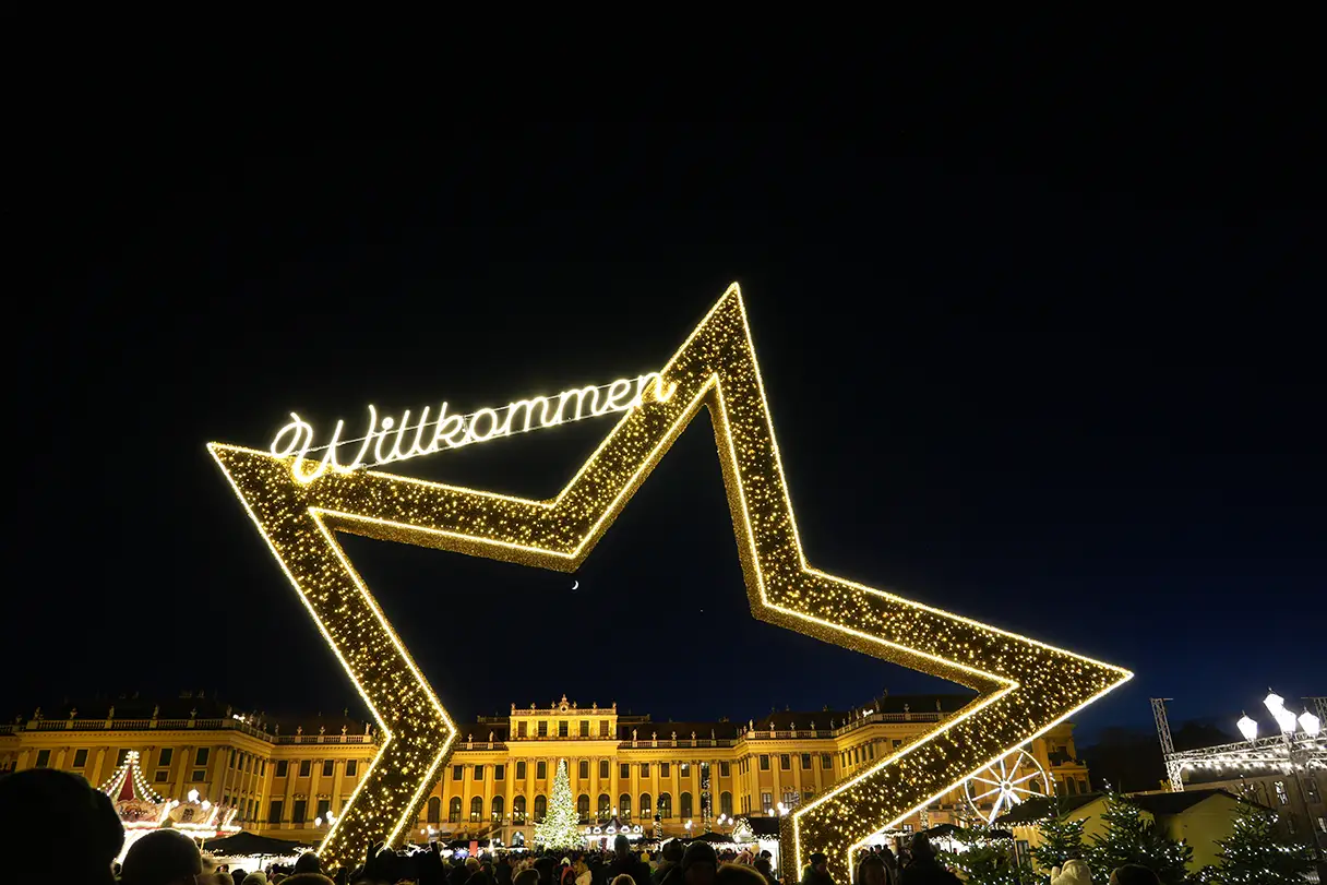 A large golden star at the entrance to the Schönbrunn Christmas Market. The golden Schönbrunn Palace can be seen in the background. A “Welcome” sign hangs from the star.