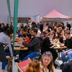 A view of the food court at the Vegan Planet Vienna trade show. Numerous visitors are standing or sitting at tables, eating or chatting. Various booths can still be seen in the background.